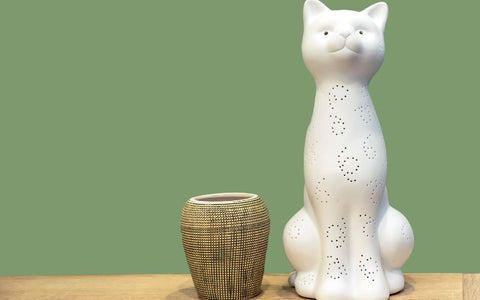 Figurine d'un chat blanc décoré de petit trou, à côté d'un petit pot. Tous les deux posés sur une table en bois. Un mur vert en fond.