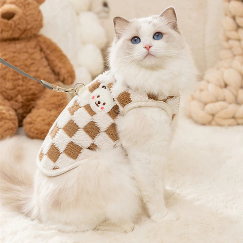 Manteau pour Chat avec Design à Carreaux porté par un chat tenu en laisse sur un tapis blanc avec des oursons en peluche en fond